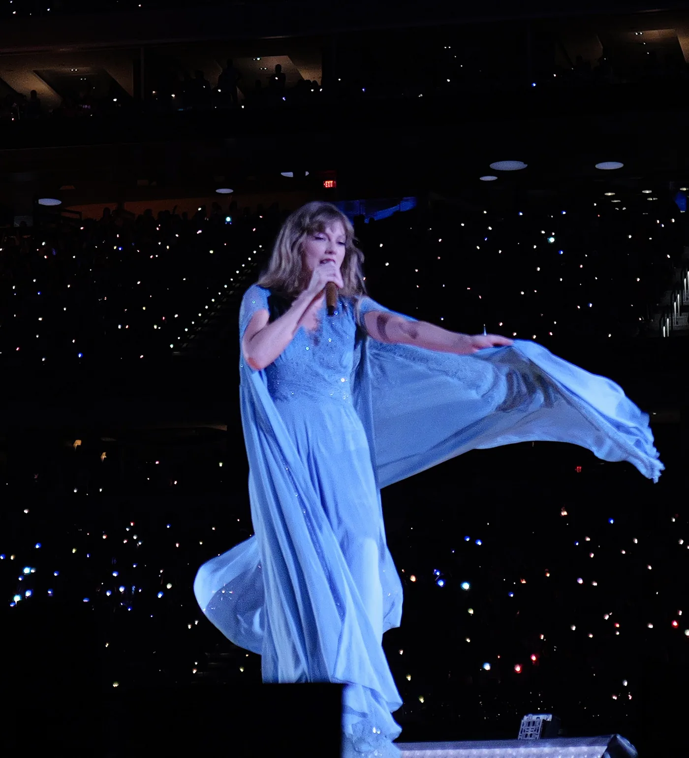 Taylor Swift singing in a flowing blue gown on the Eras Tour, a stadium of phone lights behind her